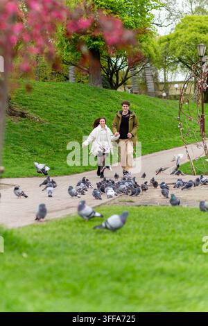 Un jeune couple heureux se tenant par la main et courant le long d'un sentier du parc tandis que des pigeons volent autour d'eux au printemps. Banque D'Images