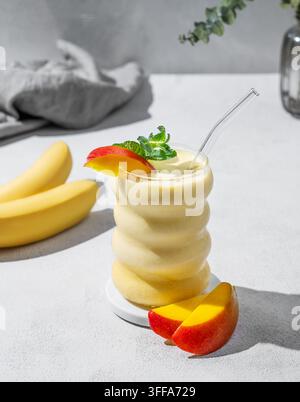 Smoothie glacé à la mangue et à la banane dans un verre sur un fond clair avec des tranches de fruits tropicaux. Concept d'alimentation saine et de régime détox. Banque D'Images