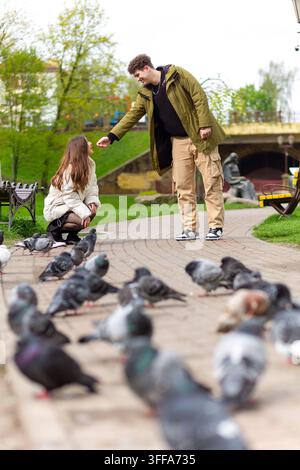 Jeune couple dans un parc urbain interagissant parmi des pigeons par une journée de printemps nuageuse avec des bancs verts et des statues Banque D'Images