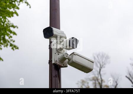 Trois caméras de surveillance montées sur un poteau, observant sous un ciel nuageux avec des feuilles d'arbres à proximité. Banque D'Images