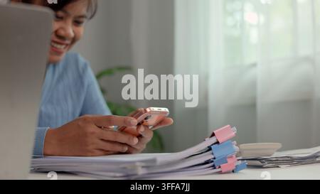 Close-up d'une femme asiatique souriante tenant un smartphone et travaillant sur un bureau avec des documents papier. Banque mobile, achats en ligne et technologie moderne Banque D'Images