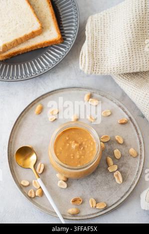 Beurre de cacahuète dans un pot en verre sur fond clair avec noix salées et toasts de pain. Concept de snack savoureux et sain pour des sandwichs sucrés pour le petit-déjeuner Banque D'Images