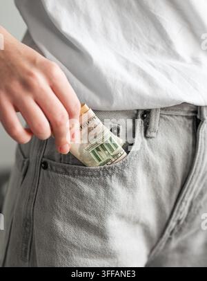 Close-up d'une main féminine mettant des billets de dollar américain roulés dans la poche avant d'un jean gris sur un fond clair à l'intérieur. Concept d'économie d'argent. Banque D'Images
