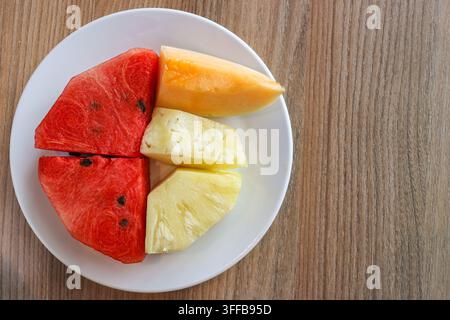 Vue de dessus de pastèque, ananas et cantaloup coupés sur une assiette blanche posée sur une table en bois avec espace libre sur le côté droit. Banque D'Images