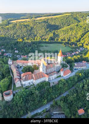 Le château de Krivoklat présente une architecture médiévale époustouflante au milieu de collines vertes luxuriantes. Cette vue aérienne capture ses tours impressionnantes et le paysage environnant lors d'un coucher de soleil doré. Banque D'Images