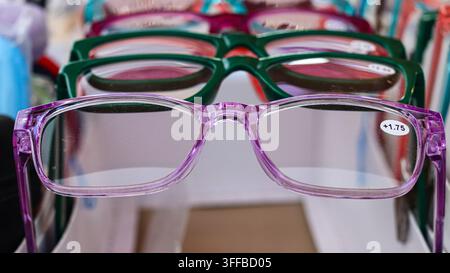 gros plan de lunettes de lecture violettes marquées +1,75 affichées devant des montures de lunettes vertes et roses dans une vitrine de vente au détail d'optique. Banque D'Images