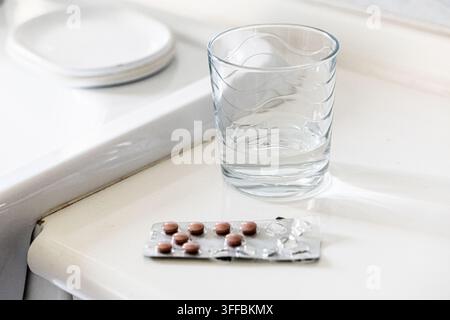 L'image montre un verre transparent contenant un peu d'eau, posé à côté d'une plaquette de petites pilules brunes sur un comptoir blanc. Banque D'Images