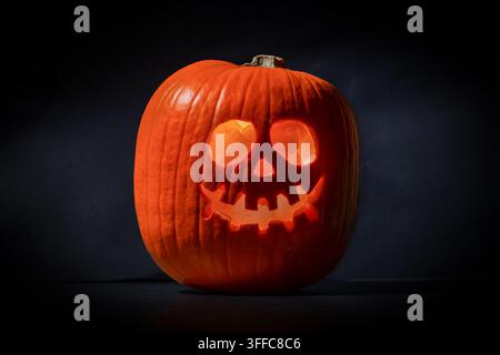 Jack-o'-lantern orange illuminé avec une tête sculptée ressemblant à un crâne, présentant des yeux ronds et des dents déchiquetées, éclairé de l'intérieur sur un fond sombre dramatique en studio Banque D'Images