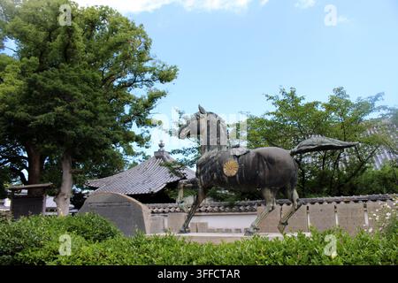 Une grande statue en bronze d'un cheval sacré orné d'une crête de chrysanthème dorée se dresse sur un piédestal en pierre devant une salle traditionnelle au toit de tuiles a Banque D'Images