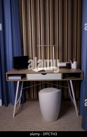 Espace de travail minimal à domicile avec ordinateur portable fermé, cahier ouvert, stylo, lampe de bureau, livres et tasse de café sur un mur rayé avec des rideaux bleus Banque D'Images