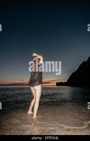 Vacances de plage au crépuscule avec une femme debout dans l'eau peu profonde levant les bras vers le ciel du soir Banque D'Images