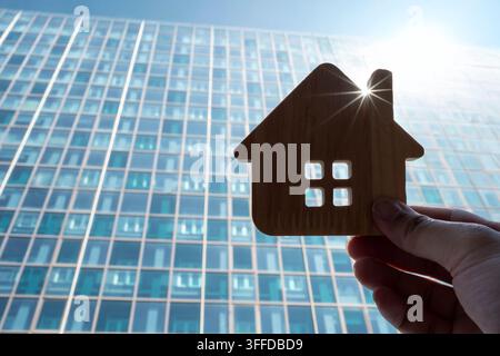 Concept d'immobilier et d'architecture moderne avec une main tenant un modèle de maison en bois devant un immeuble en verre de type gratte-ciel avec éclat de soleil pour la ville Banque D'Images