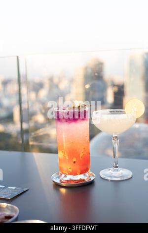 Deux cocktails élégants posés sur une table sombre dans un bar rooftop en hauteur – une boisson ombre vibrante stratifiée en rouge, rose et violet avec une garniture florale, un Banque D'Images