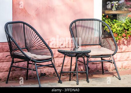 Siège extérieur lumineux d'un café avec des chaises en osier noires confortables dotées de coussins moelleux et une table basse en verre assortie le long d'un trottoir. Banque D'Images