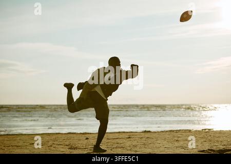 Homme noir actif lançant un ballon de football américain sur une plage de sable Banque D'Images