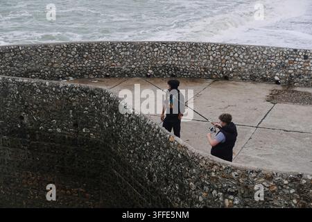 Deux jeunes hommes pêchant avec des cannes à pêche depuis un môle en pierre dans l'océan agité à Brighton, en Angleterre. Banque D'Images