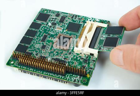 Une main connecte un petit module mémoire à une carte électronique verte dans un atelier. Le décor montre diverses pièces électroniques. Banque D'Images
