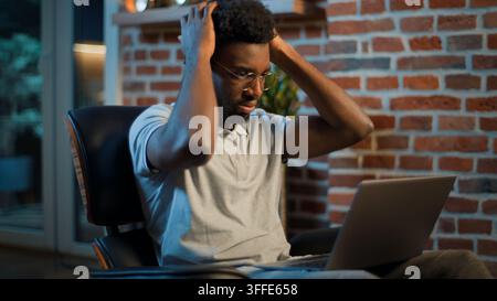 Homme d'affaires afro-américain stressé et inquiet avec ordinateur portable se sentant contrarié dans un bureau du soir, homme d'affaires surmené, travailleur masculin avec erreur informatique Banque D'Images