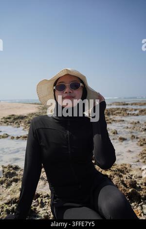 Une femme asiatique portant un hijab noir, des lunettes de soleil et un chapeau de soleil est assise détendue sur une plage tropicale rocheuse avec des formations coralliennes et une eau de mer claire sous un bri Banque D'Images
