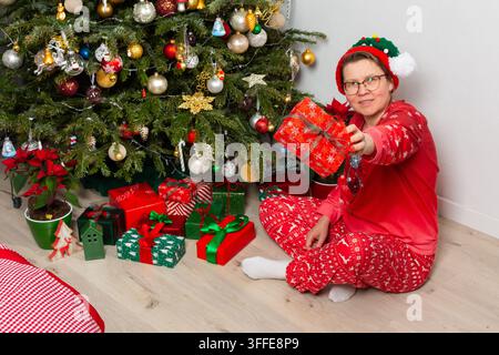 Le jour de Noël, une femme de 52 ans est assise par terre près d'un sapin de Noël. Elle sourit tout en tenant des cadeaux emballés et entourée de décorations dans son salon confortable Banque D'Images