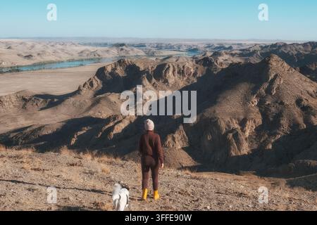 Une femme avec son chien se tient sur une colline surplombant le canyon rocheux et la rivière Ili dans une vaste vallée aride sous un ciel bleu. Ce paysage de style de vie est p Banque D'Images