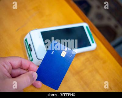 Un gros plan d'une main tenant une carte de crédit ou de débit en plastique moderne contre un terminal de paiement sans contact (POS) blanc et élégant pour compléter Banque D'Images