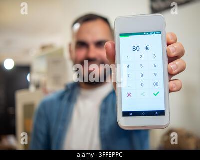 Close-up d'un homme barbu non identifiable tenant un lecteur de carte POS moderne avec un montant de 80 € prêt pour un paiement sans contact. Banque D'Images