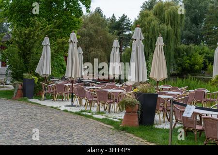 Une rangée de grands parapluies crème fermés s'élève au-dessus de rangées de tables et de chaises en osier rose vides sur une terrasse pavée près d'un étang. Banque D'Images