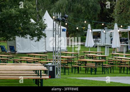Des rangées de bancs et de tables en bois vides sont disposées sur une pelouse sous des guirlandes lumineuses, près de tentes blanches et de parasols fermés. Banque D'Images
