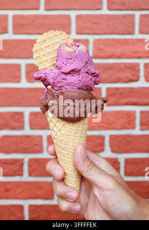 Main tenant une boule double de yaourt à la framboise et une boule de gelato au chocolat noir dans un cornet contre un mur de briques Banque D'Images