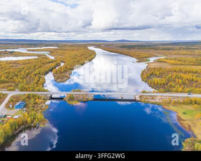Vue aérienne d'un pont routier enjambant une rivière miroitante reflétant le ciel et les nuages, entourée d'une nature sauvage dorée de l'automne dans le nord de la Suède Banque D'Images