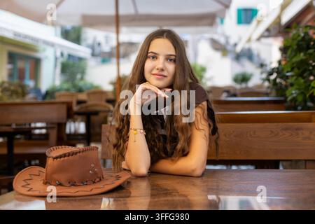 Jeune femme assise à une table de café avec un chapeau marron. Jeune femme aux longs cheveux bruns assise à une table de café en bois, posant son menton sur sa main à côté d'un Banque D'Images