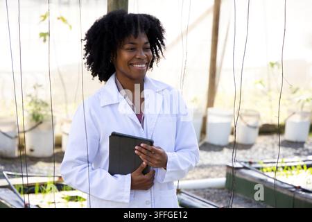 Femme africaine d'âge moyen debout dans une serre, portant une blouse de laboratoire blanche et tenant une tablette Banque D'Images
