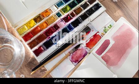 Espace de travail d'un artiste aquarelliste. Verre d'eau, un pinceau, un mouchoir taché et une palette d'aquarelle sur une table blanche. Gros plan du processus créatif. Banque D'Images