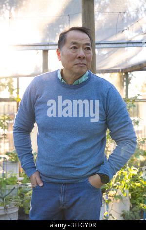 Homme asiatique âgé debout dans une serre parmi des plantes en pot près d'un poteau en bois, portant un pull bleu Banque D'Images
