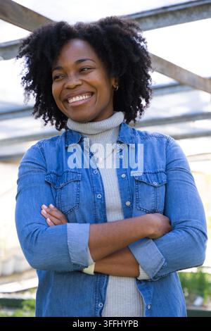 Femme afro-américaine d'âge moyen debout sous une pergola métallique, souriante, portant une chemise en jean et un collier Banque D'Images
