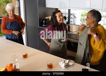 Différentes amies âgées riant en tabliers en préparant des boissons glacées près d'une citrouille sur l'îlot de cuisine Banque D'Images