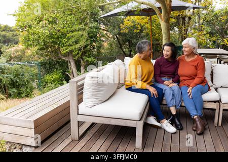 Diverses amies féminines assises sur un canapé d'angle en bois rembourré sur la terrasse sous un parasol, bavardant Banque D'Images