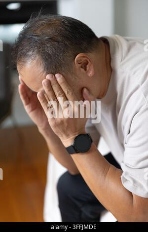 Homme asiatique âgé assis sur une chaise à la maison, se penchant en avant en couvrant son visage, montre visible, plancher en bois Banque D'Images