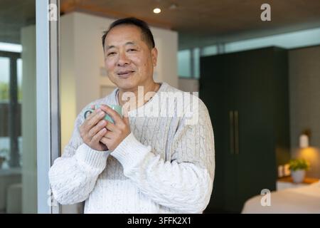 Homme asiatique âgé debout dans une chambre tenant une tasse bleu-vert, portant un pull en tricot crème Banque D'Images