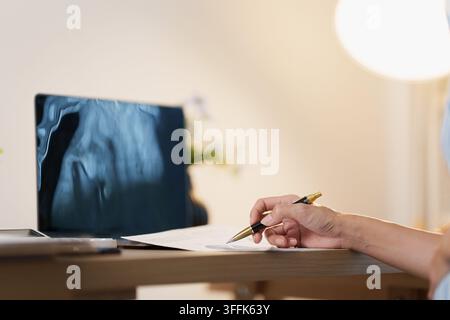 Main coupée écrivant sur un document près d'un ordinateur portable affichant une radiographie médicale pour un rapport de santé Banque D'Images