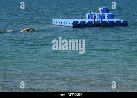 Un nageur solitaire traverse l'eau claire du lac à côté d'une plateforme de natation flottante, entouré de ondulations par une journée d'été ensoleillée et chaude. Banque D'Images