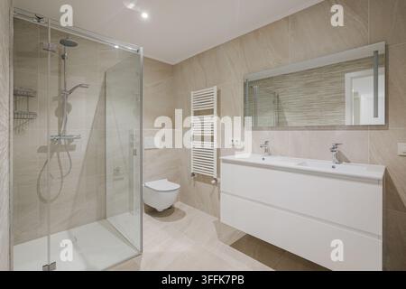Intérieur de salle de bain moderne avec douche en verre, lavabo double blanc, murs carrelés beige, toilettes murales, miroir rectangulaire, accessoires chromés et Banque D'Images