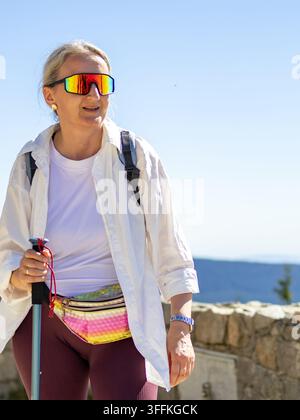 Femme souriant joyeusement au sommet de Szrenica portant des lunettes de soleil avec des bâtons de trekking et un sac à dos. Concept de réalisation joyeuse et d'arrivée réussie au sommet Banque D'Images