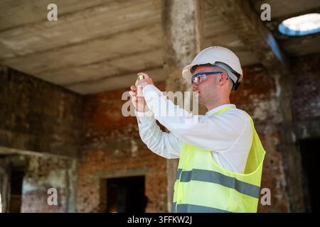 Professionnel de la construction utilisant un smartphone sur un chantier inachevé tout en portant un équipement de sécurité. Gestion numérique des chantiers inspection à distance Banque D'Images