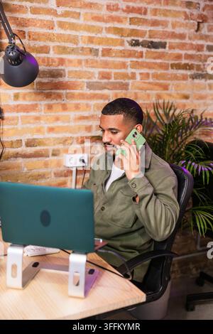 Homme latin concentré travaillant sur un ordinateur portable et parlant au téléphone dans un bureau moderne Banque D'Images