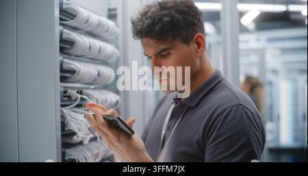 Technicien de centre de données masculin vérifie le câblage Ethernet et les étiquettes des ports physiques par rapport à la documentation réseau sur smartphone. Collègue féminine effectue Banque D'Images