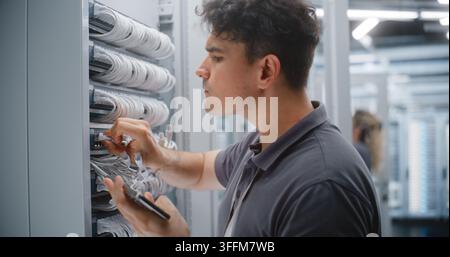 Technicien de centre de données masculin vérifie le câblage Ethernet et les étiquettes des ports physiques par rapport à la documentation réseau sur smartphone. Collègue féminine effectue Banque D'Images