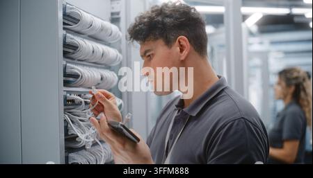 Technicien de centre de données masculin vérifie le câblage Ethernet et les étiquettes des ports physiques par rapport à la documentation réseau sur smartphone. Collègue féminine effectue Banque D'Images