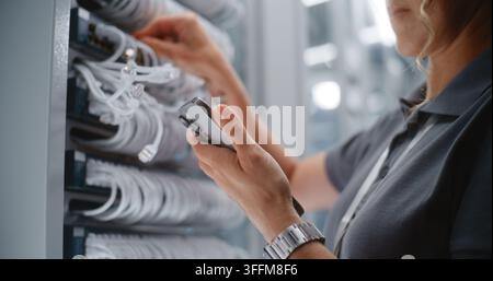 Technicienne de réseau féminine effectuant un audit des ports physiques à côté d’un rack de serveurs, utilisant un smartphone pour vérifier l’identification des câbles et les enregistrements du réseau. Concept Banque D'Images